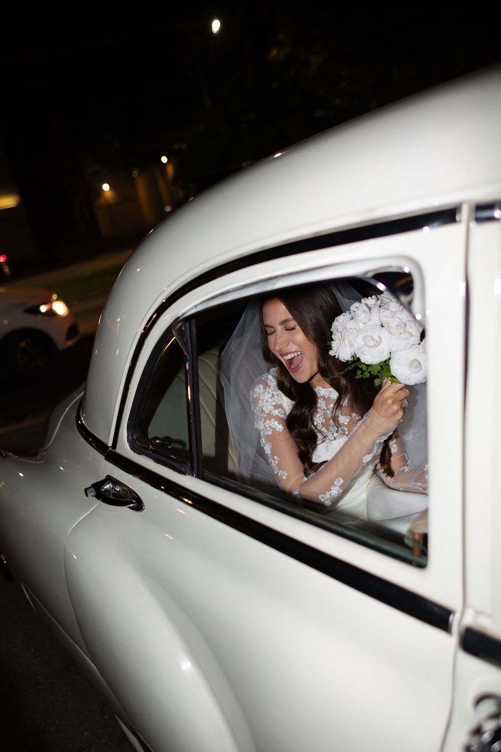 A joyful moment captured through the passenger window of a vintage white car during a wedding celebration.