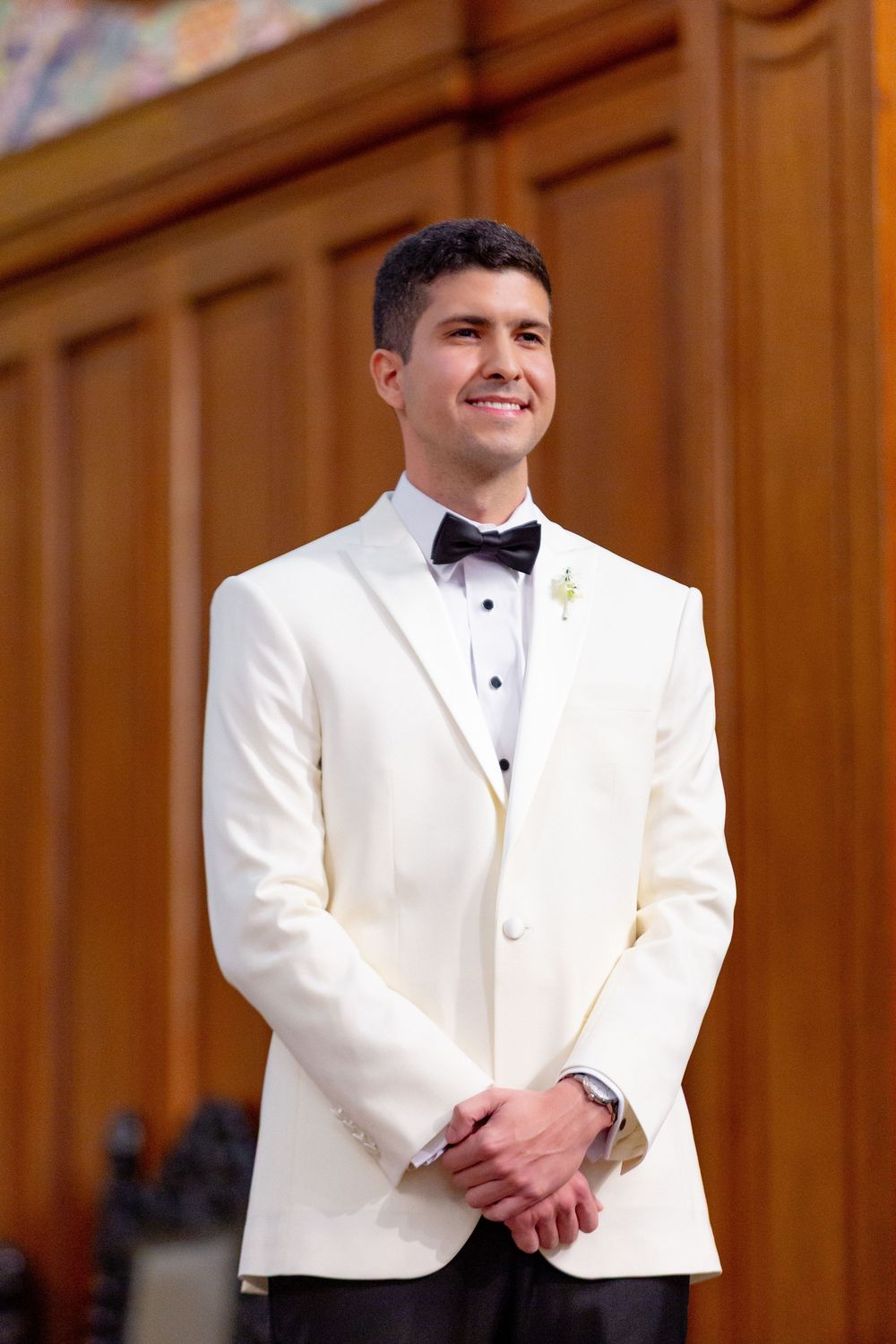 A person in a white dinner jacket and black bow tie smiles during a formal event in a wood-paneled room.