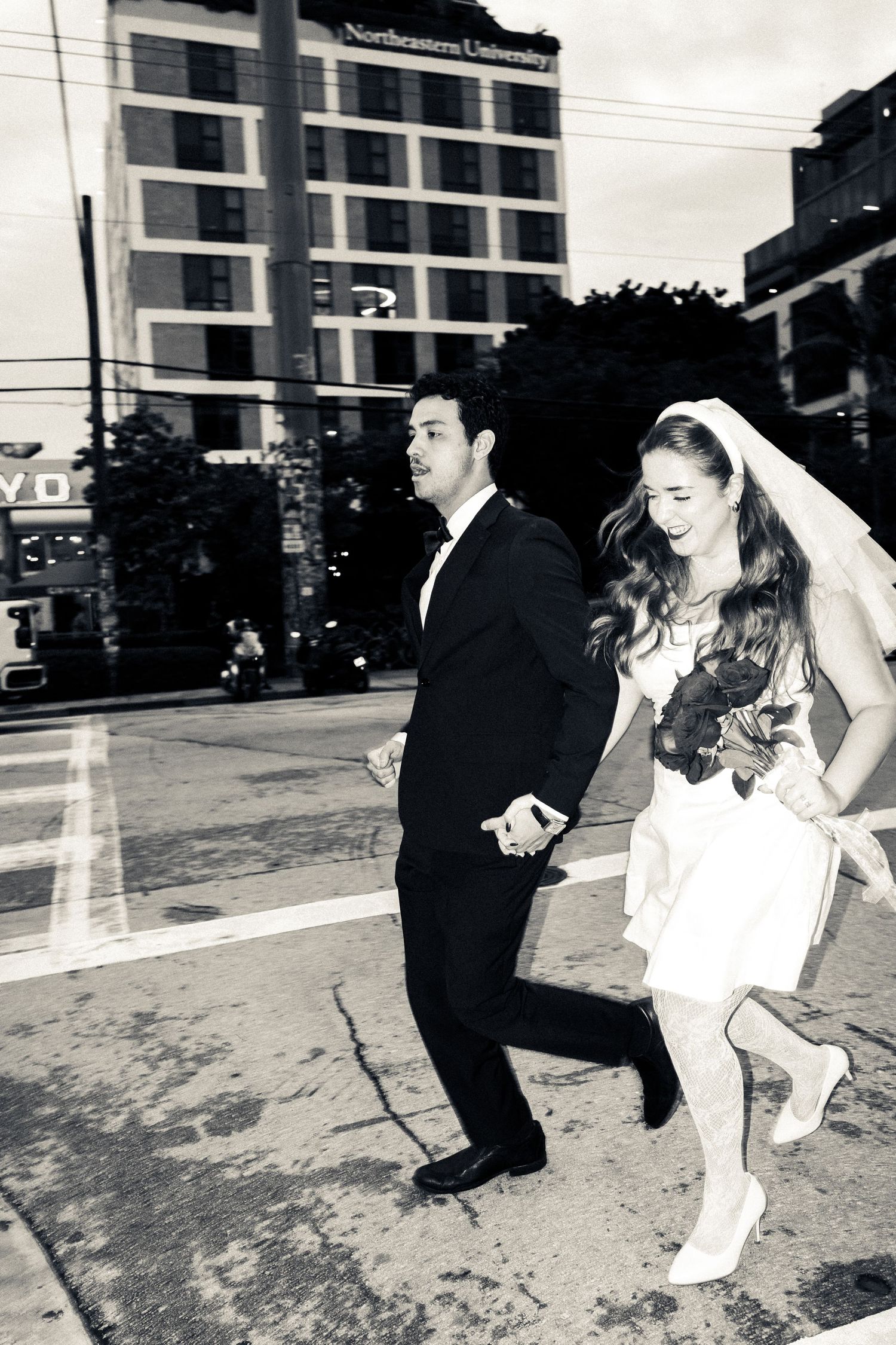 A couple in wedding attire run playfully through an urban street with a tall building in the background.
