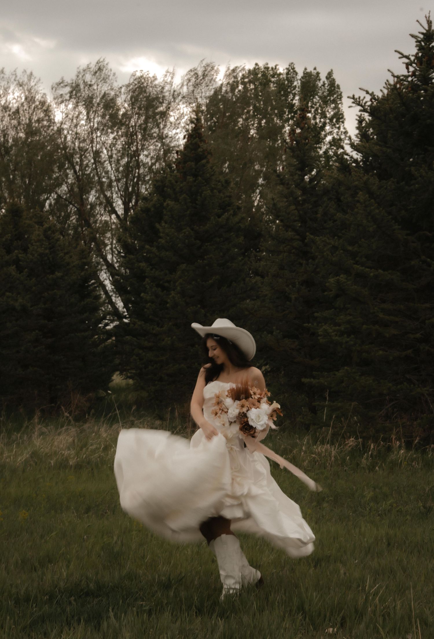 A wedding dress billows dramatically in the wind as a couple poses against an evergreen forest backdrop at dusk.