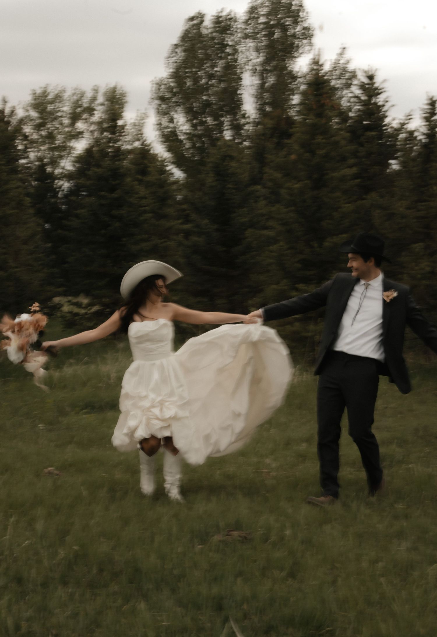 A joyful wedding couple dances together in a grassy field with evergreen trees in the background.