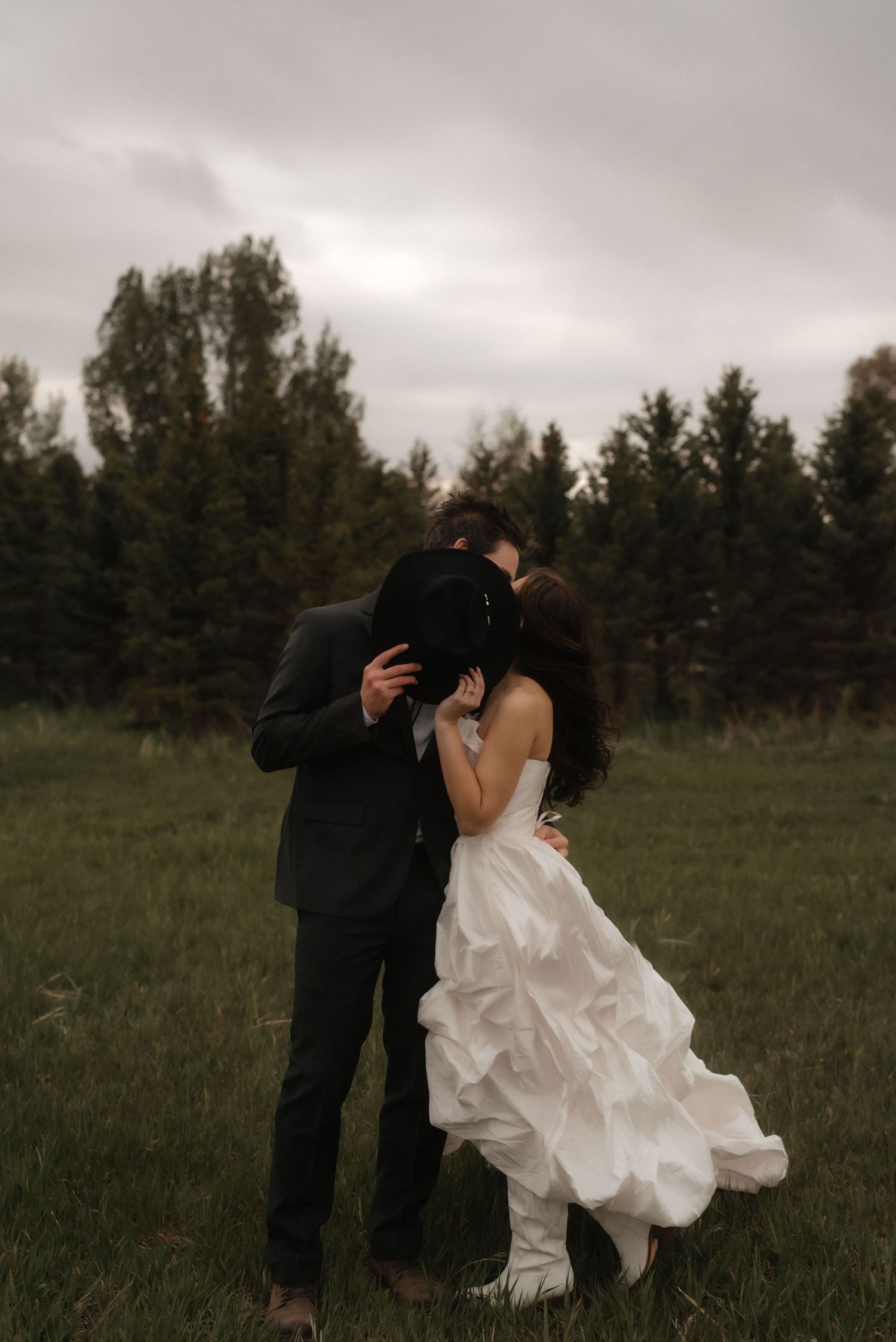 A romantic couple dances together in a grassy field with evergreen trees and moody cloudy skies in the background.