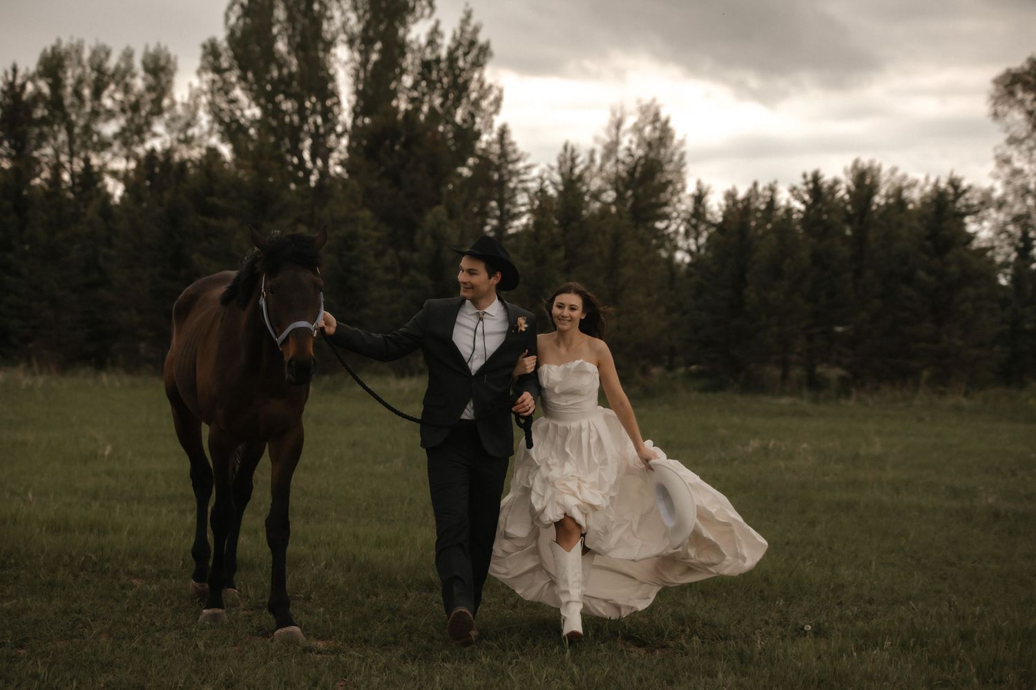 A romantic evening wedding shoot in a rustic field features a couple with horses under dramatic sunset skies.