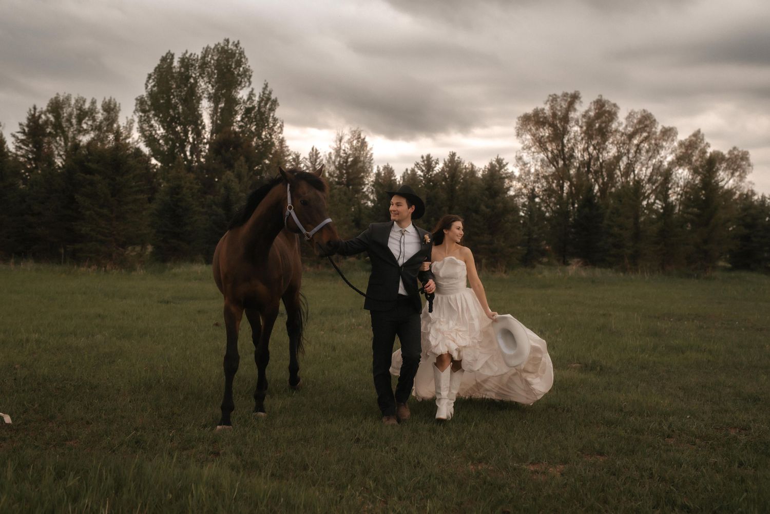 Wedding couple poses in a field with horses during sunset under dramatic cloudy skies.