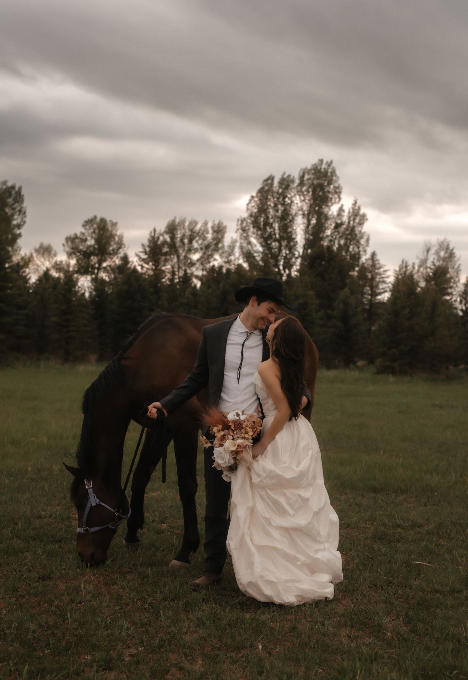 A couple in wedding attire stands in a grassy field with their brown horse under dramatic cloudy skies.