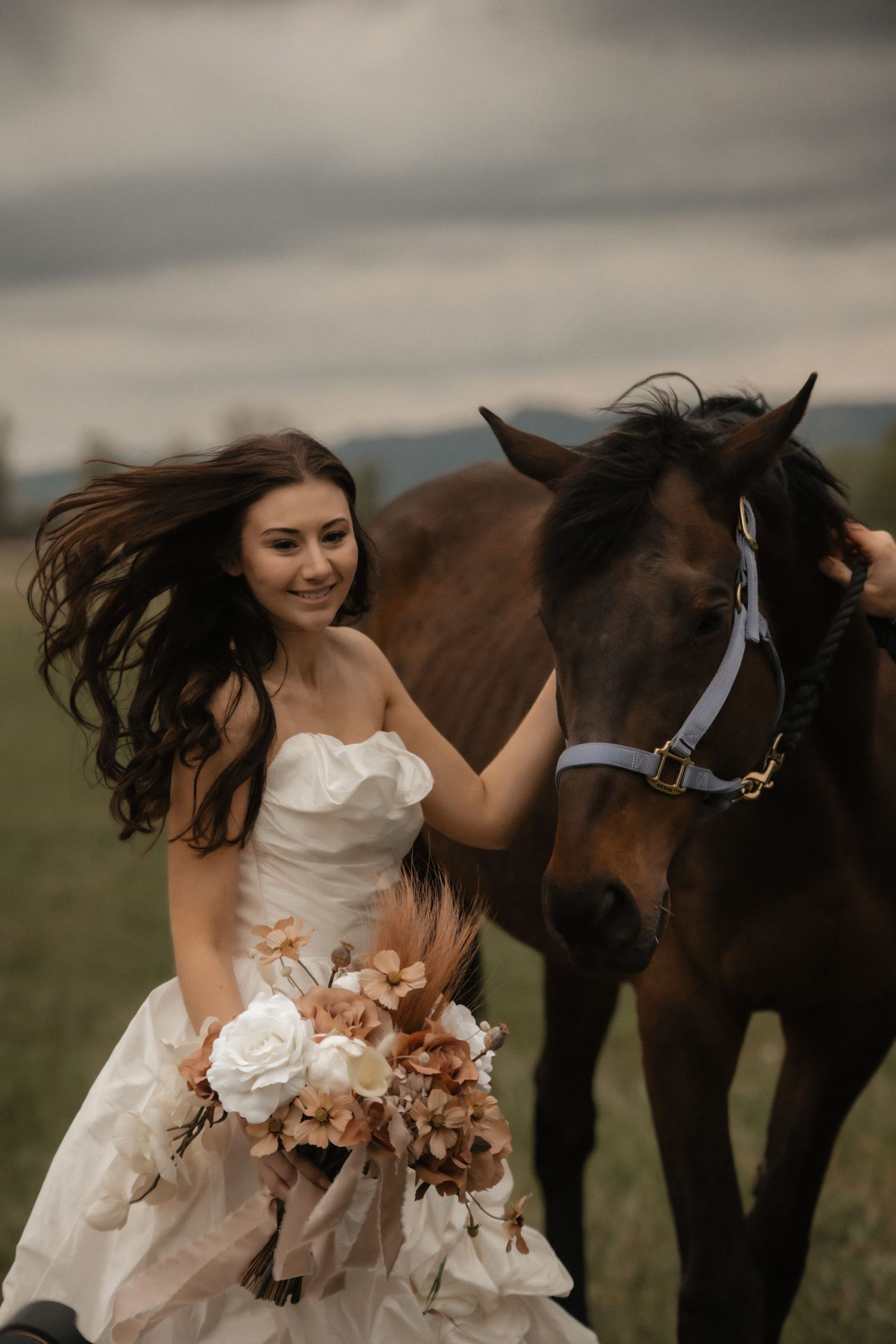 A bride in a white dress holding a peach bouquet stands next to a brown horse in a field.