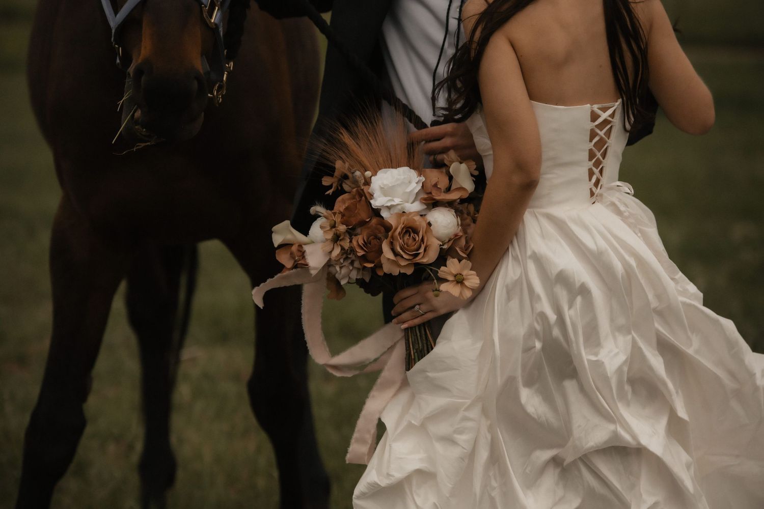 Two people in wedding attire stand close together outdoors with bouquet of pink and peach roses.