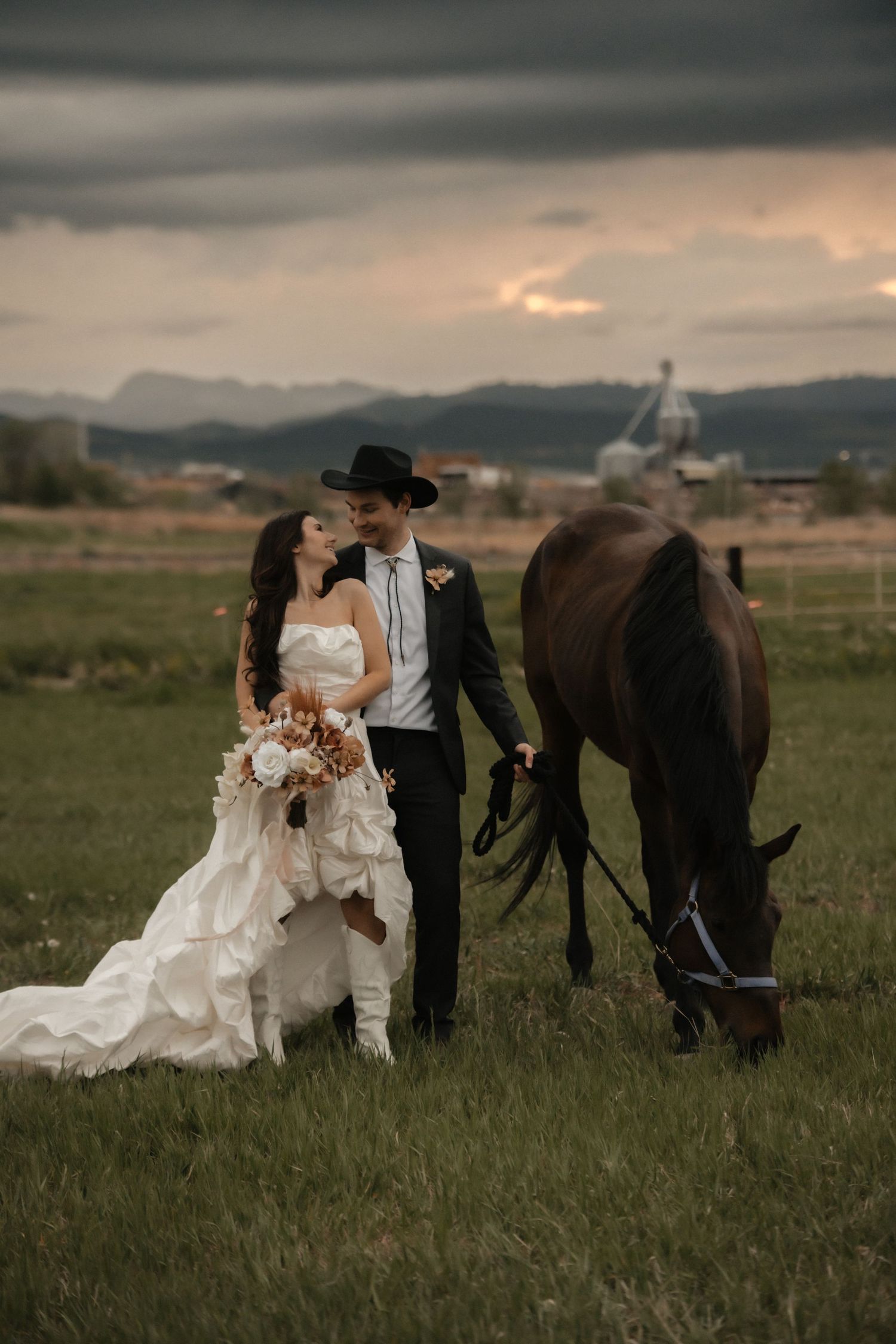 A romantic country wedding scene with a couple standing in a field alongside a grazing brown horse at sunset.
