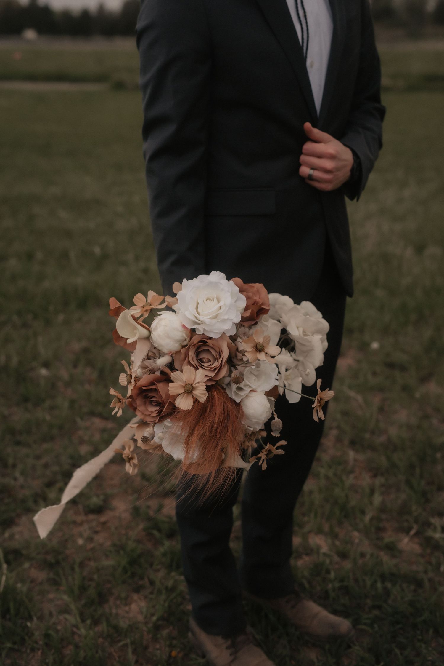 A person in a dark suit holds a romantic bridal bouquet with white and dusty rose flowers against a grassy field backdrop.