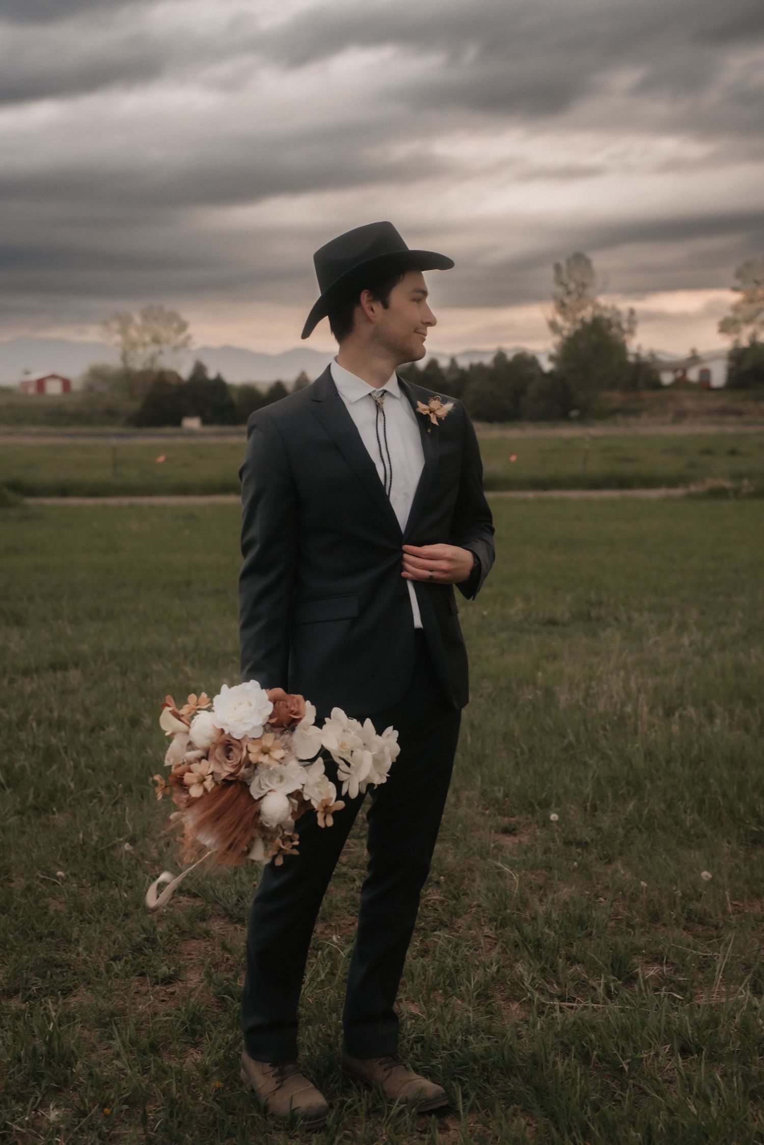 A formal figure in a black suit and cowboy hat stands in a field at sunset holding a floral bouquet.