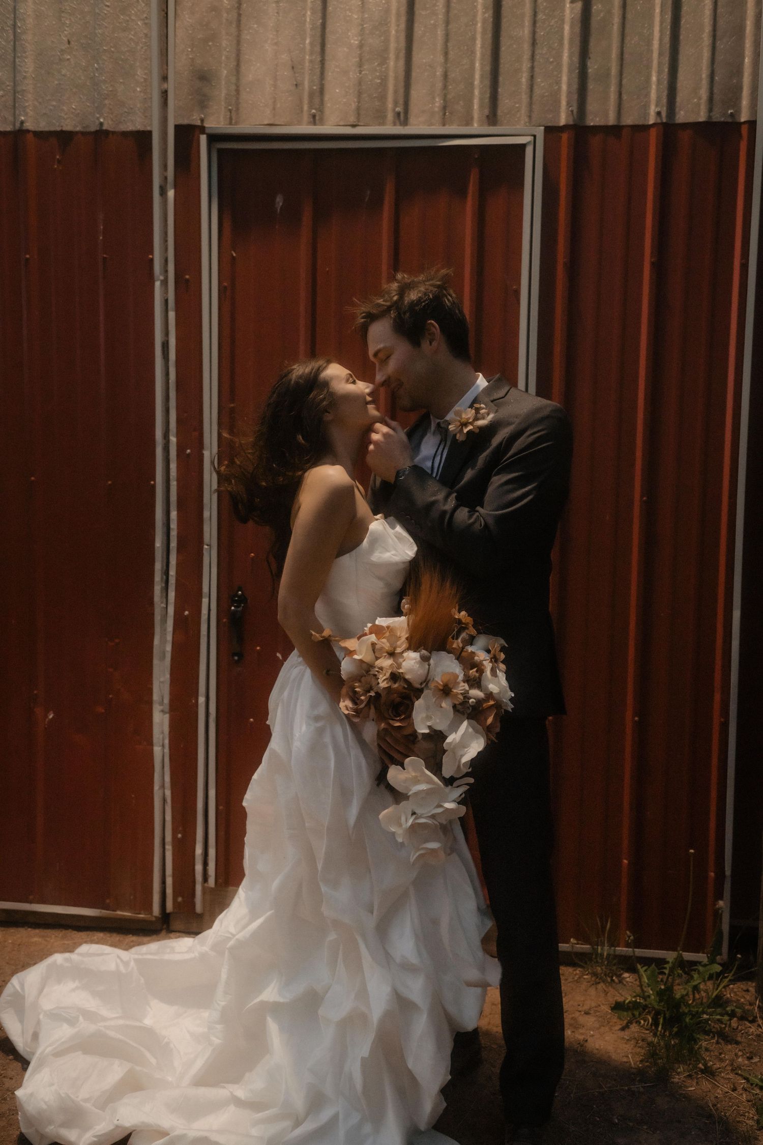 A romantic couple in wedding attire shares kisses against a rustic red barn backdrop.