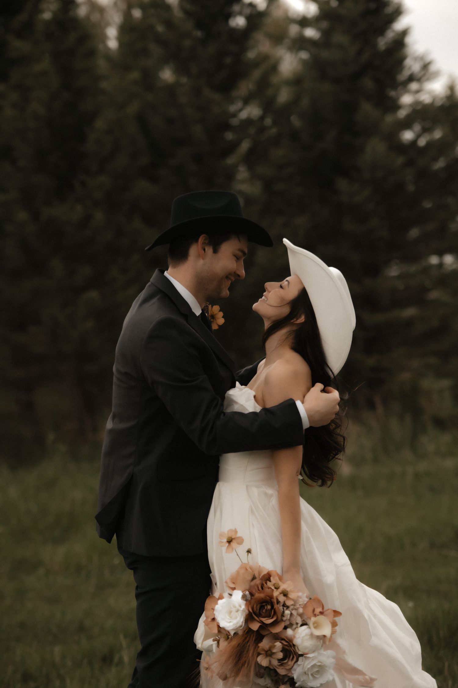 A romantic wedding couple embraces in a wooded field at sunset with the bride holding a bouquet of autumn-colored flowers.