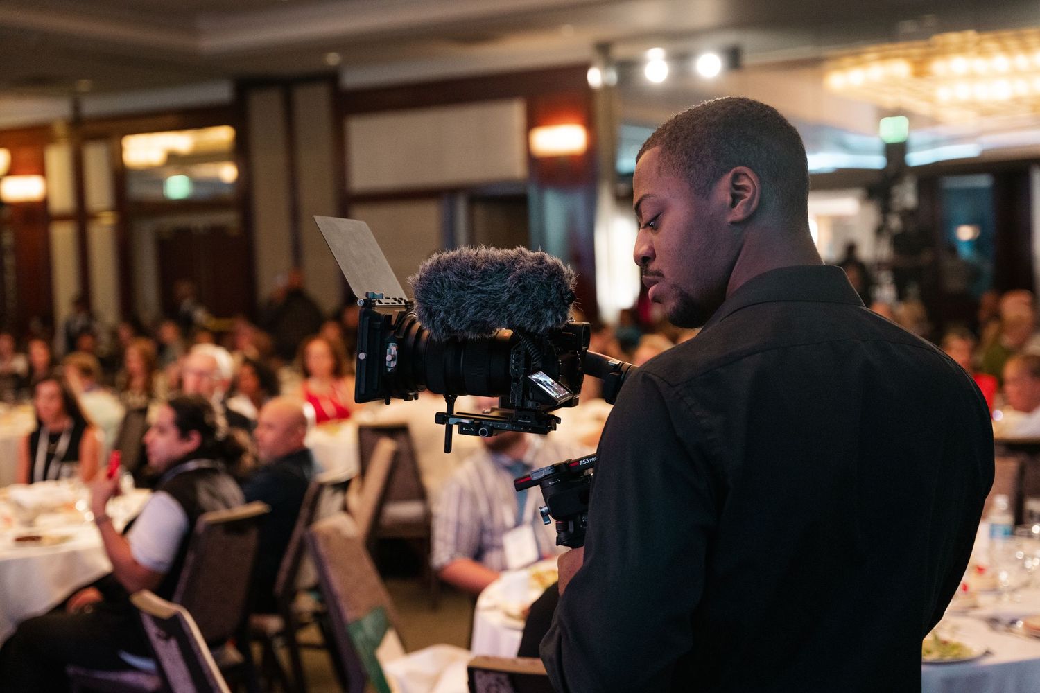 A videographer operates a professional camera at an indoor event with tables and guests in the background.