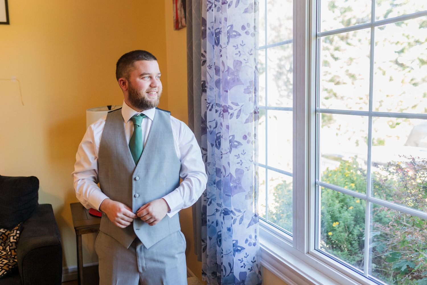Standing by a sunny window in formal attire including a vest and tie while getting ready for a special occasion.