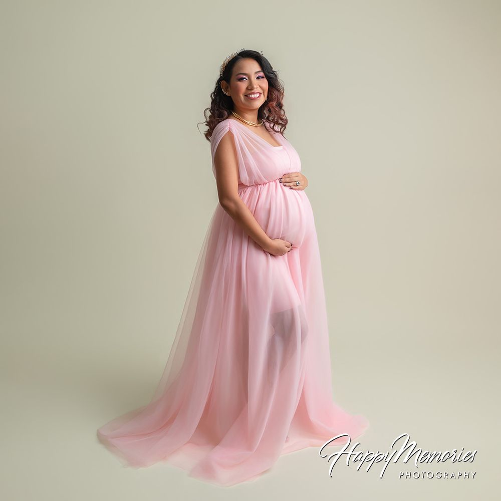Smiling pregnant woman in flowing pink tulle maternity gown poses gracefully in a professional studio photo.