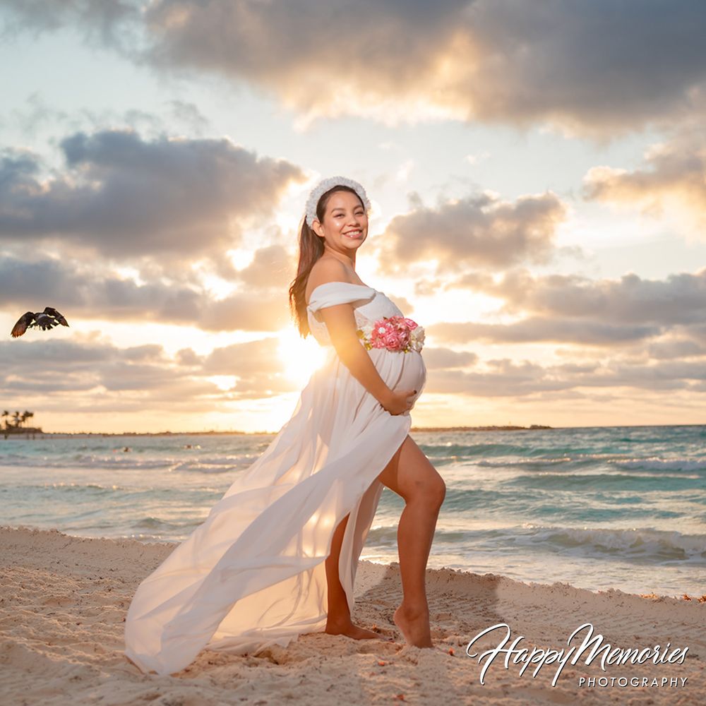 Pregnant woman in white off-shoulder gown holding pink flowers on a beach at sunset with ocean waves behind her.