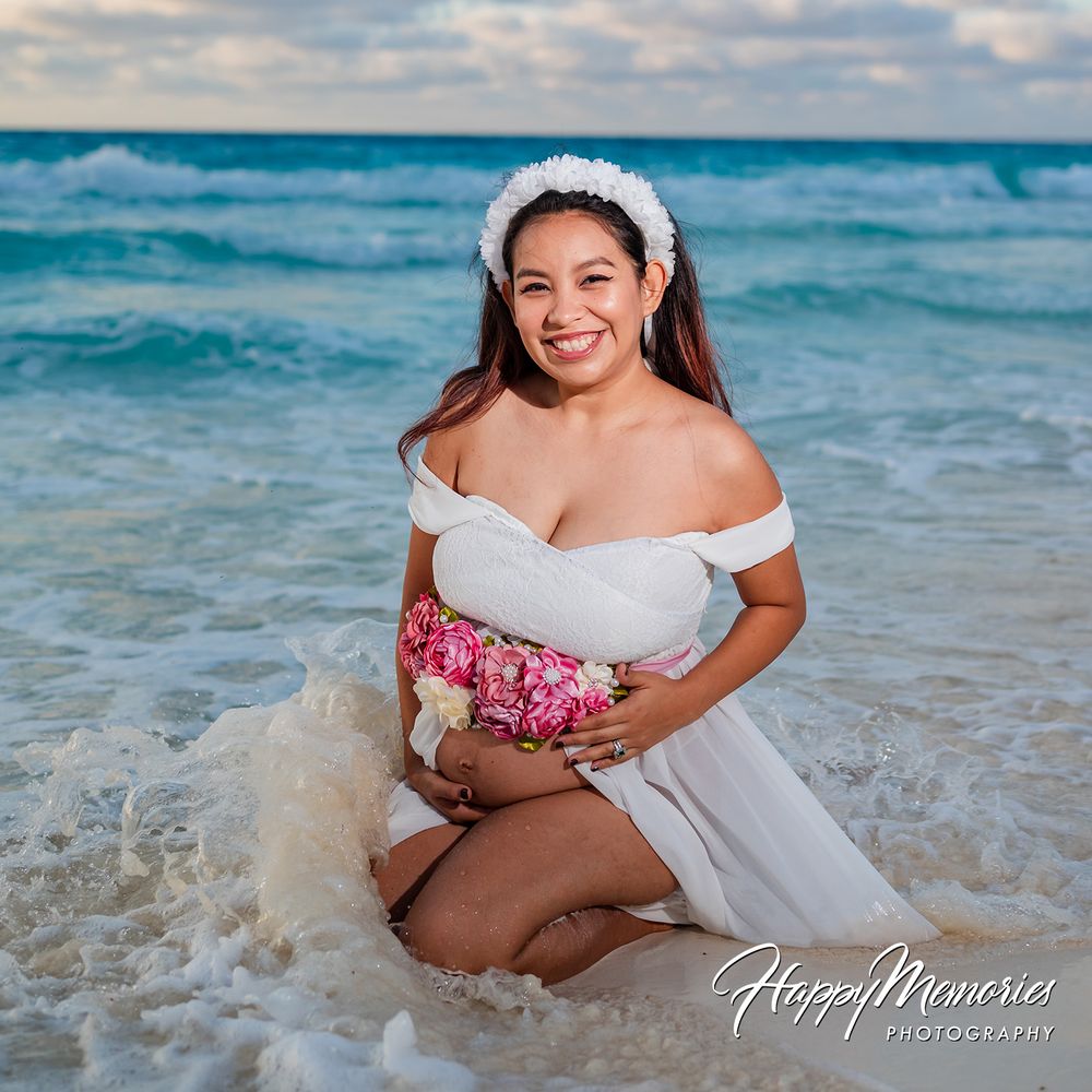Pregnant woman in white off-shoulder gown kneeling in ocean waves, smiling, wearing floral headband, holding baby bump.
