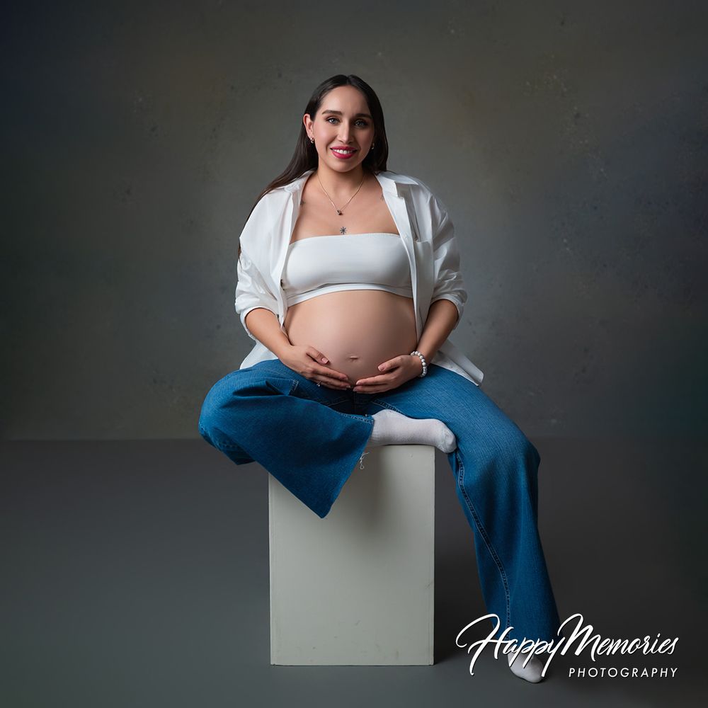 Pregnant woman in white crop top and blue pants cradling baby bump, seated on white cube for maternity photography session.