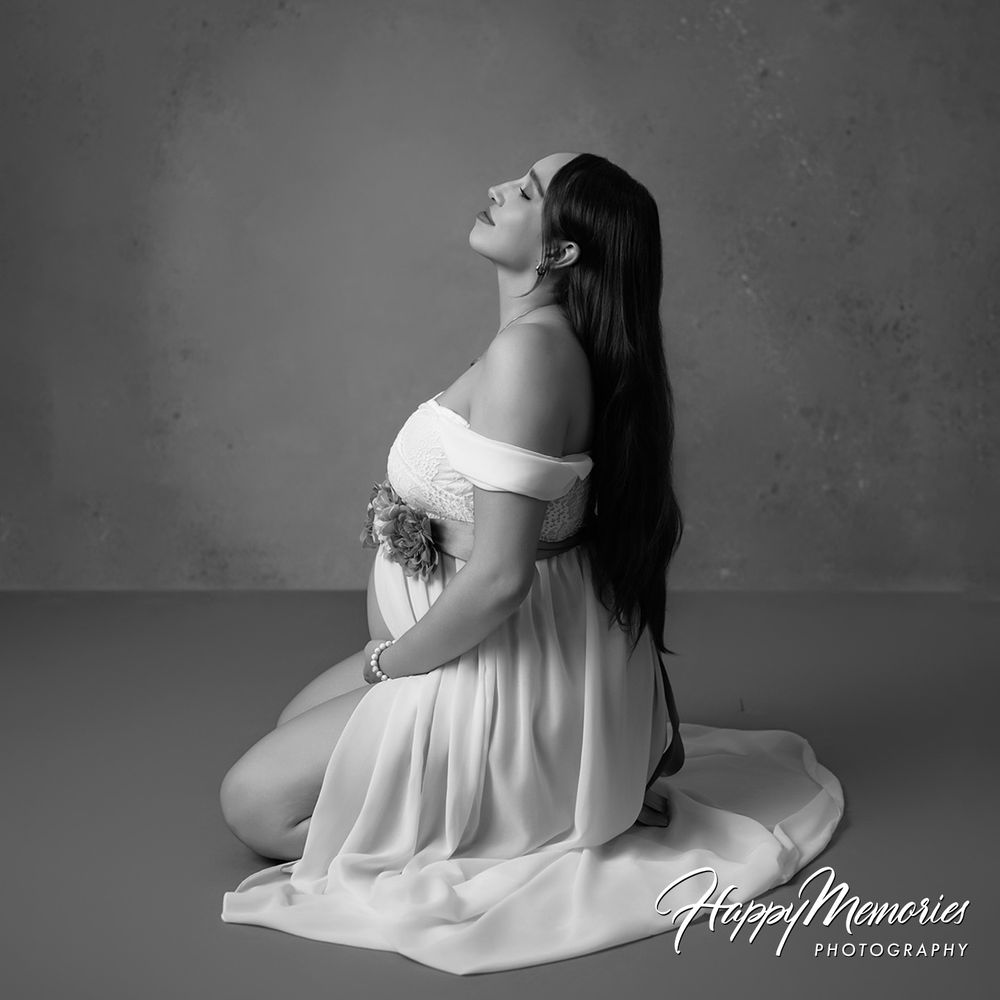 Elegant black and white maternity photo of pregnant woman kneeling in flowing white gown.