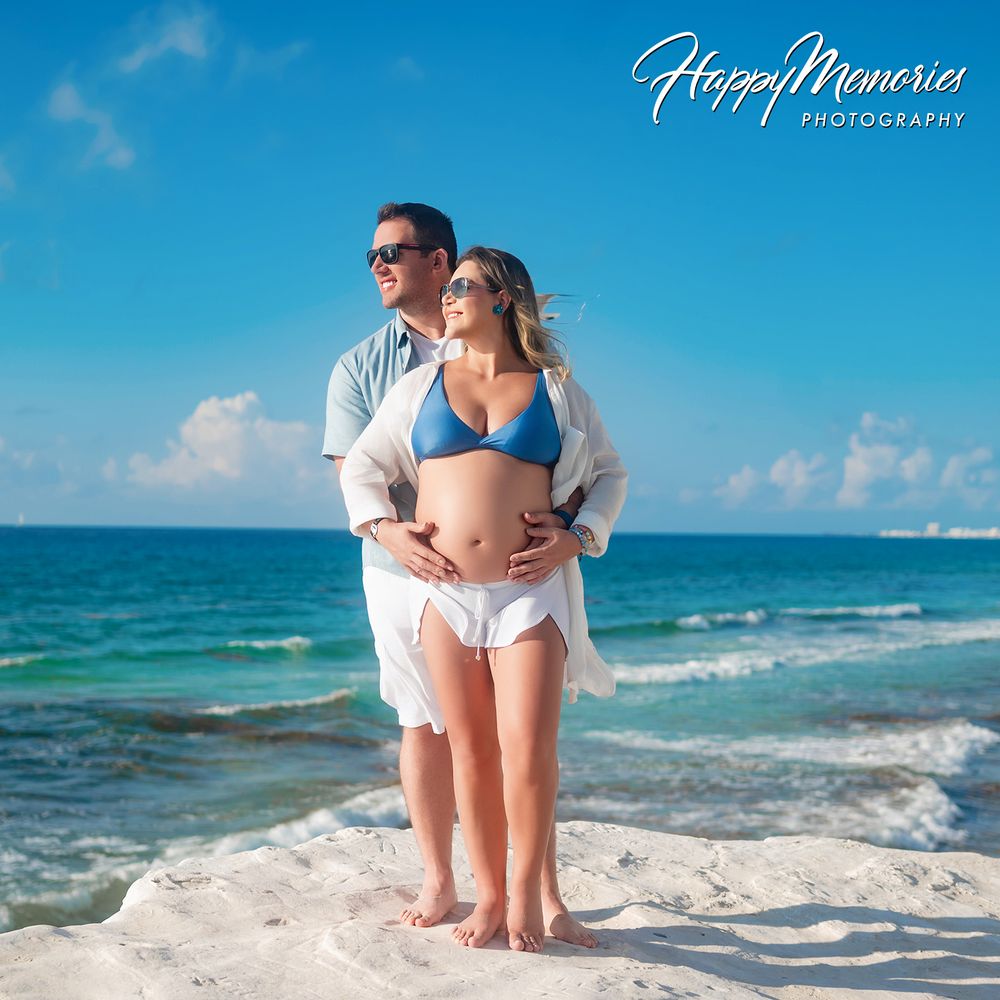 Pregnant couple posing on a tropical beach, woman in blue bikini, man embracing her from behind, turquoise ocean backdrop.