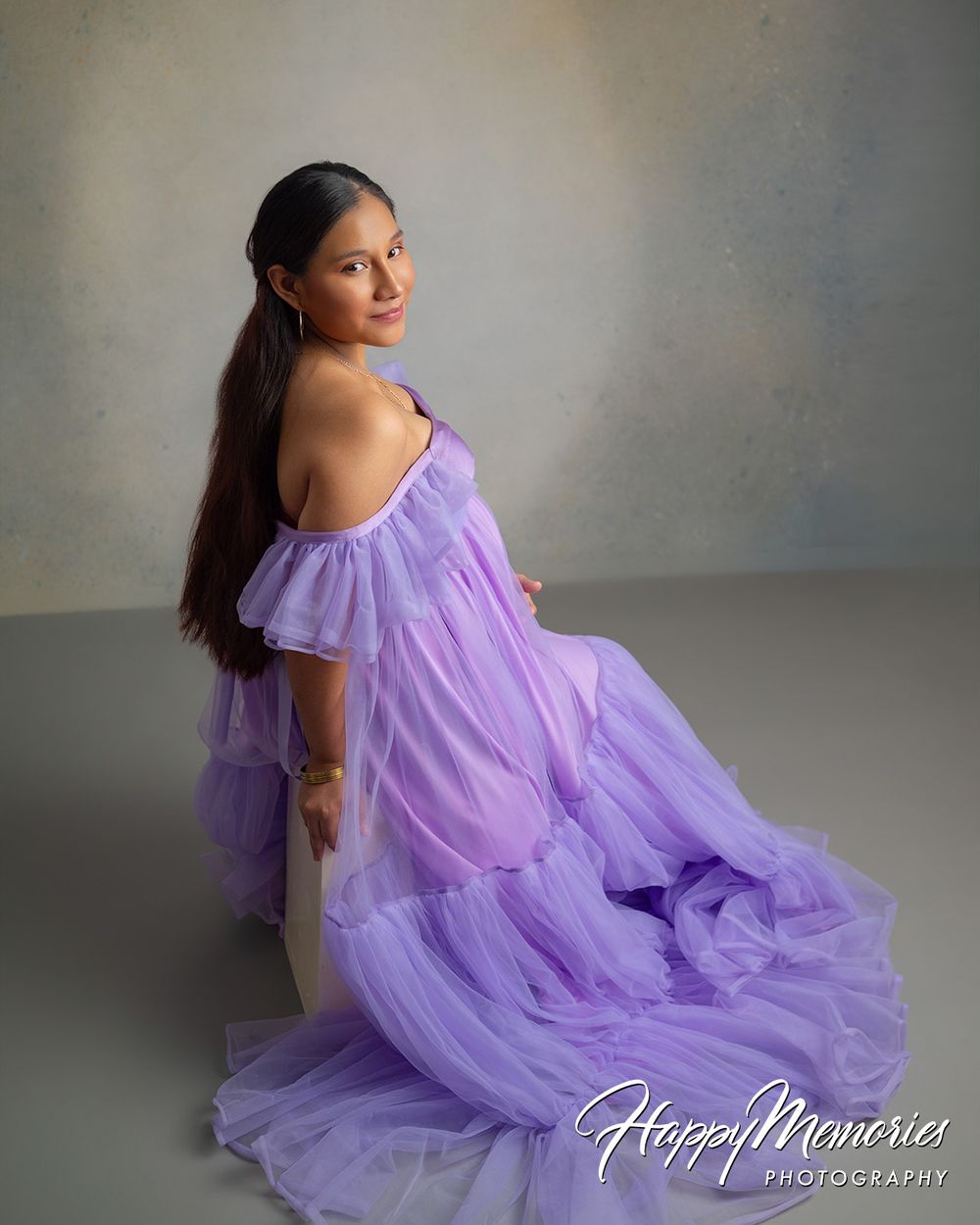 Pregnant woman in flowing lavender tulle gown poses gracefully in a professional maternity photo studio shoot.
