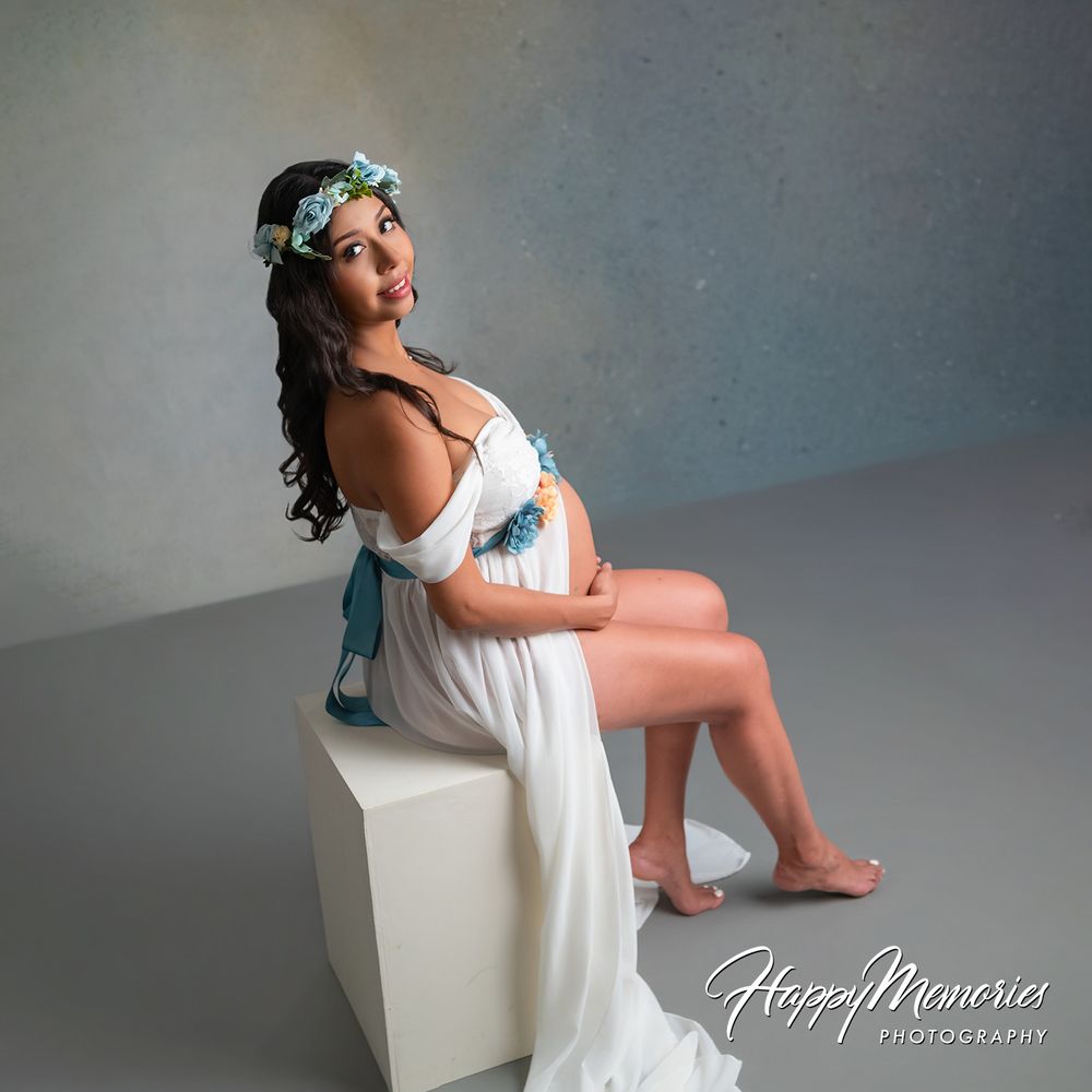 Pregnant woman in white off-shoulder maternity gown with floral crown poses gracefully on white cube in professional studio.