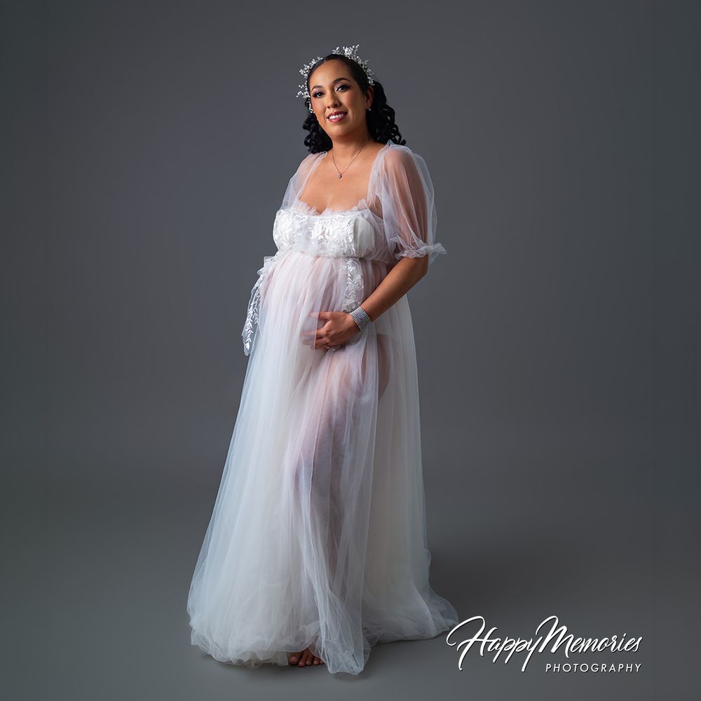 Pregnant woman in elegant white tulle maternity gown with floral crown stands beautifully in professional photography studio.
