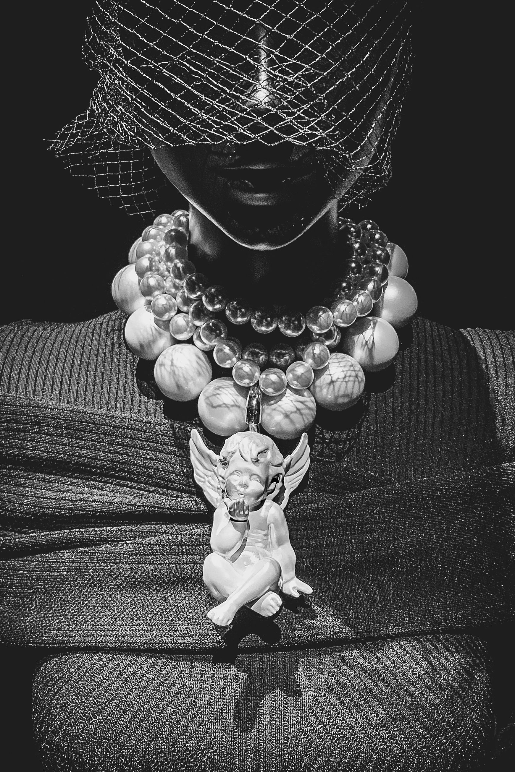 Dramatic black and white photo showing ornate beaded necklace with cherub pendant and delicate mesh veil on dark background.