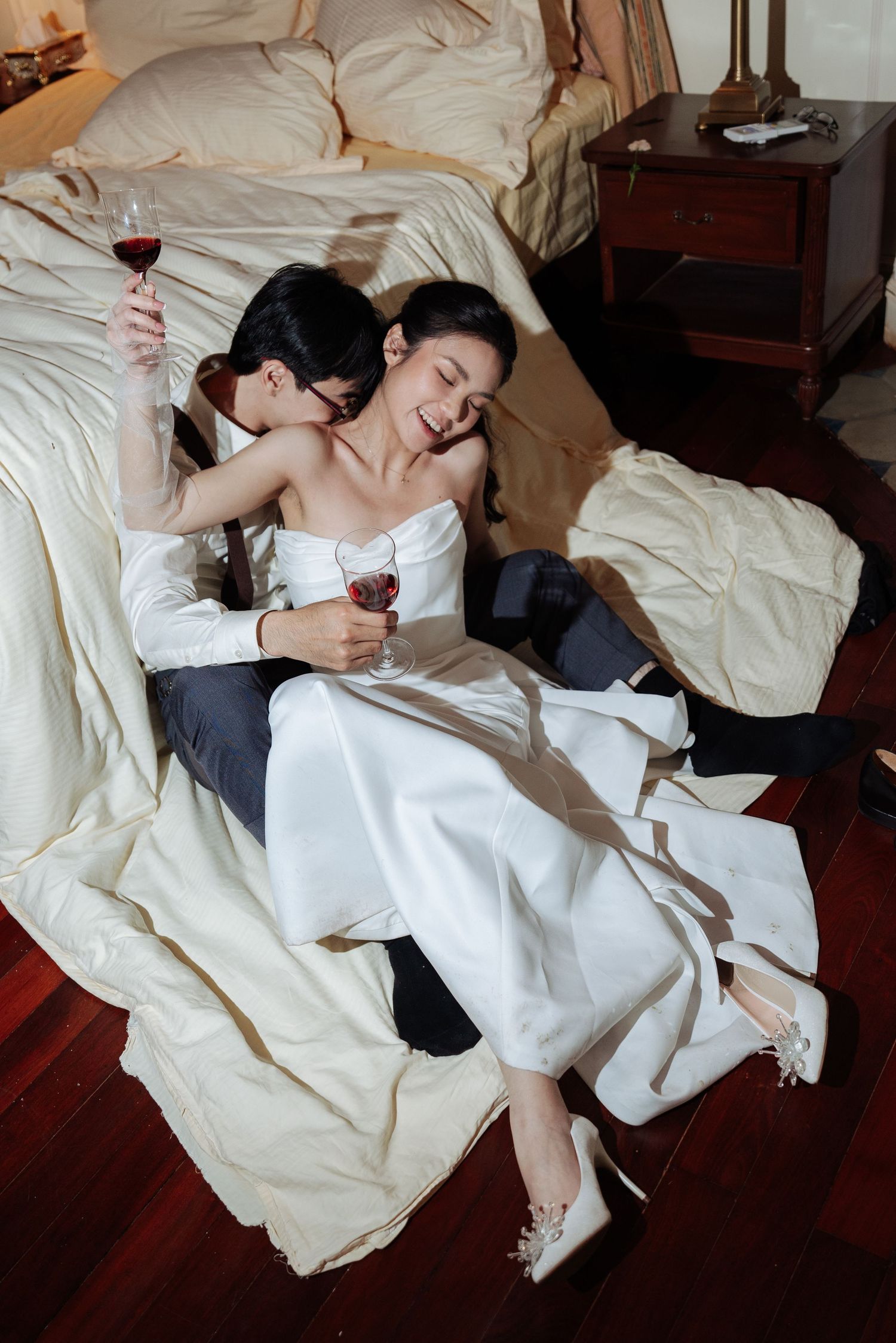 Bride and groom sit on bedroom floor laughing and drinking red wine together in wedding attire.