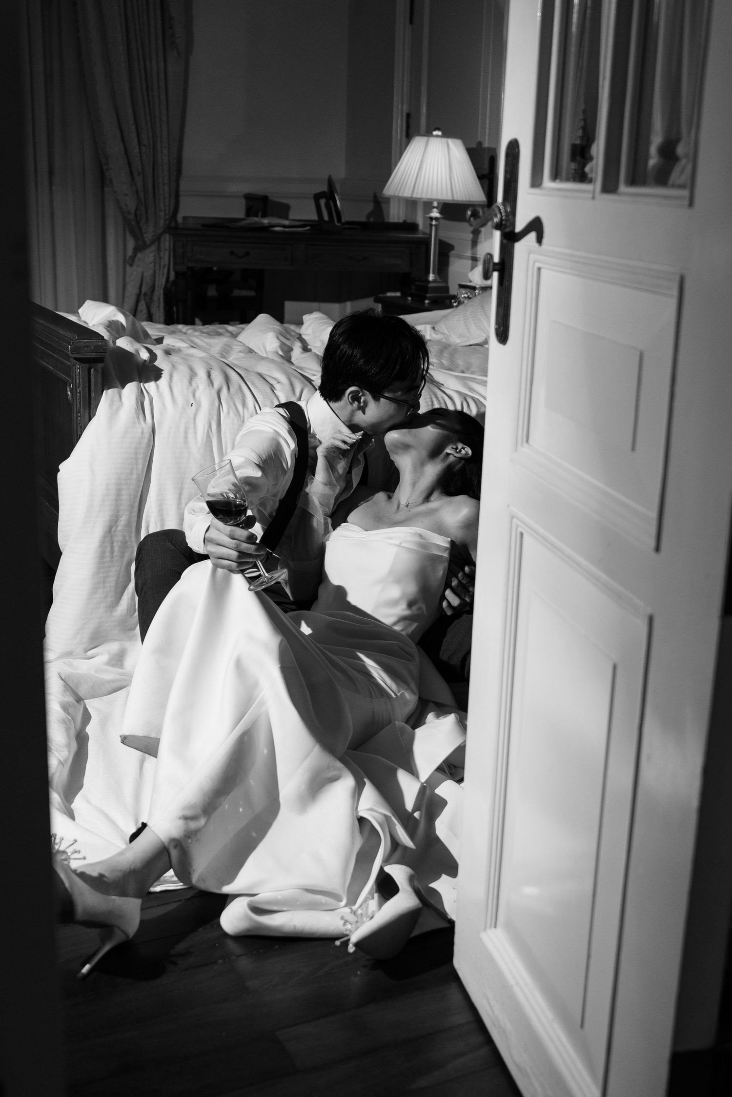 Black and white photo of couple kissing on floor beside a bed viewed through an open doorway.