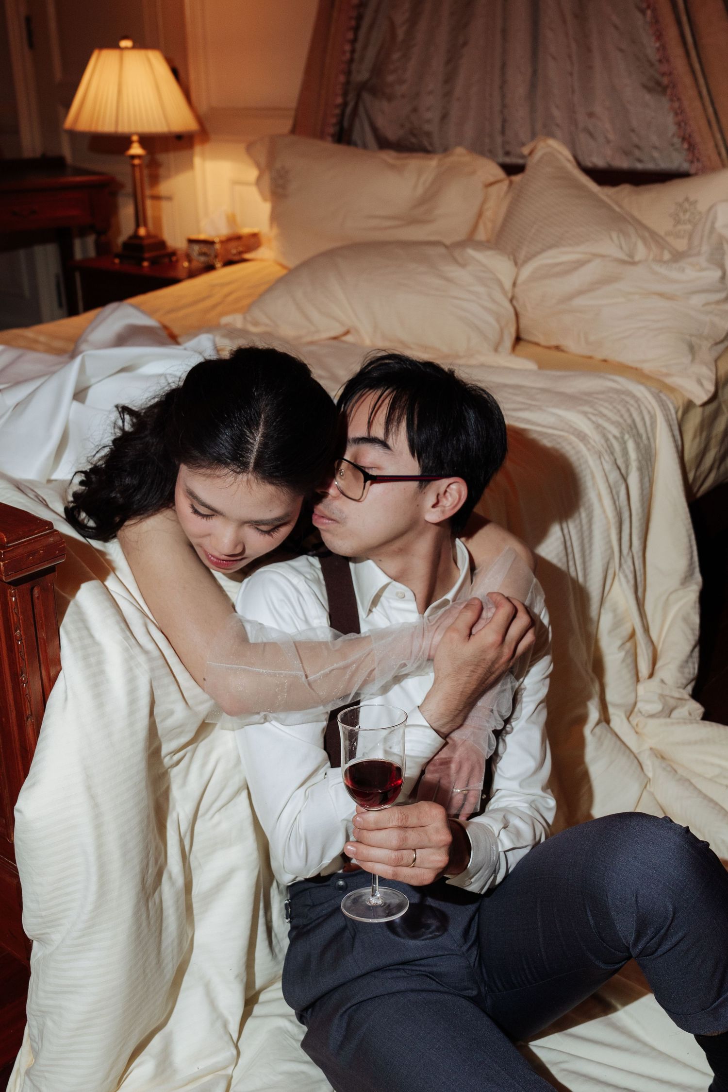 Bride rests her head on grooms shoulder as they sit together on bedroom floor holding red wine.