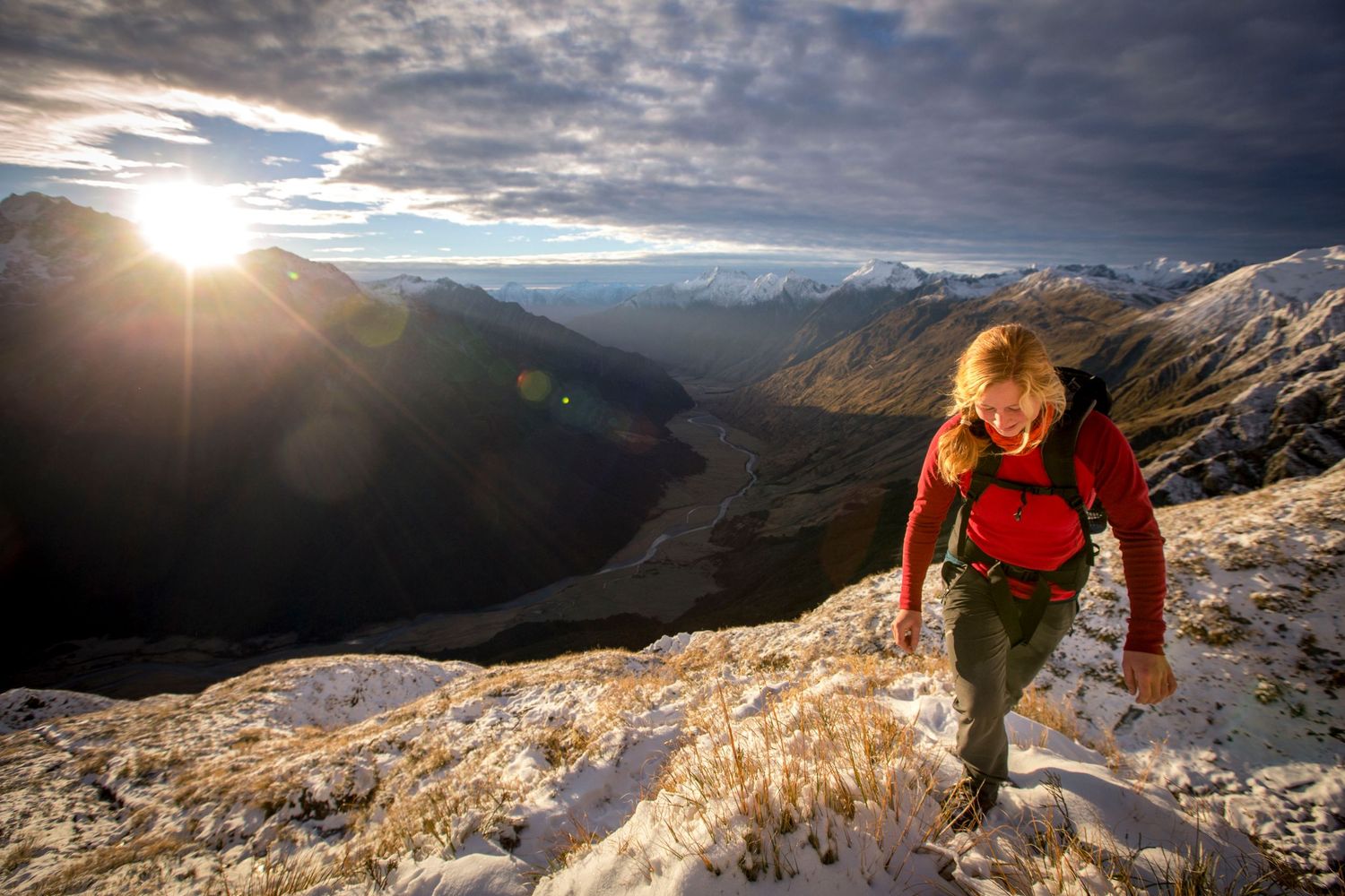 Female hiker climbing snowy mountain ridge with dramatic valley and sunset in background.