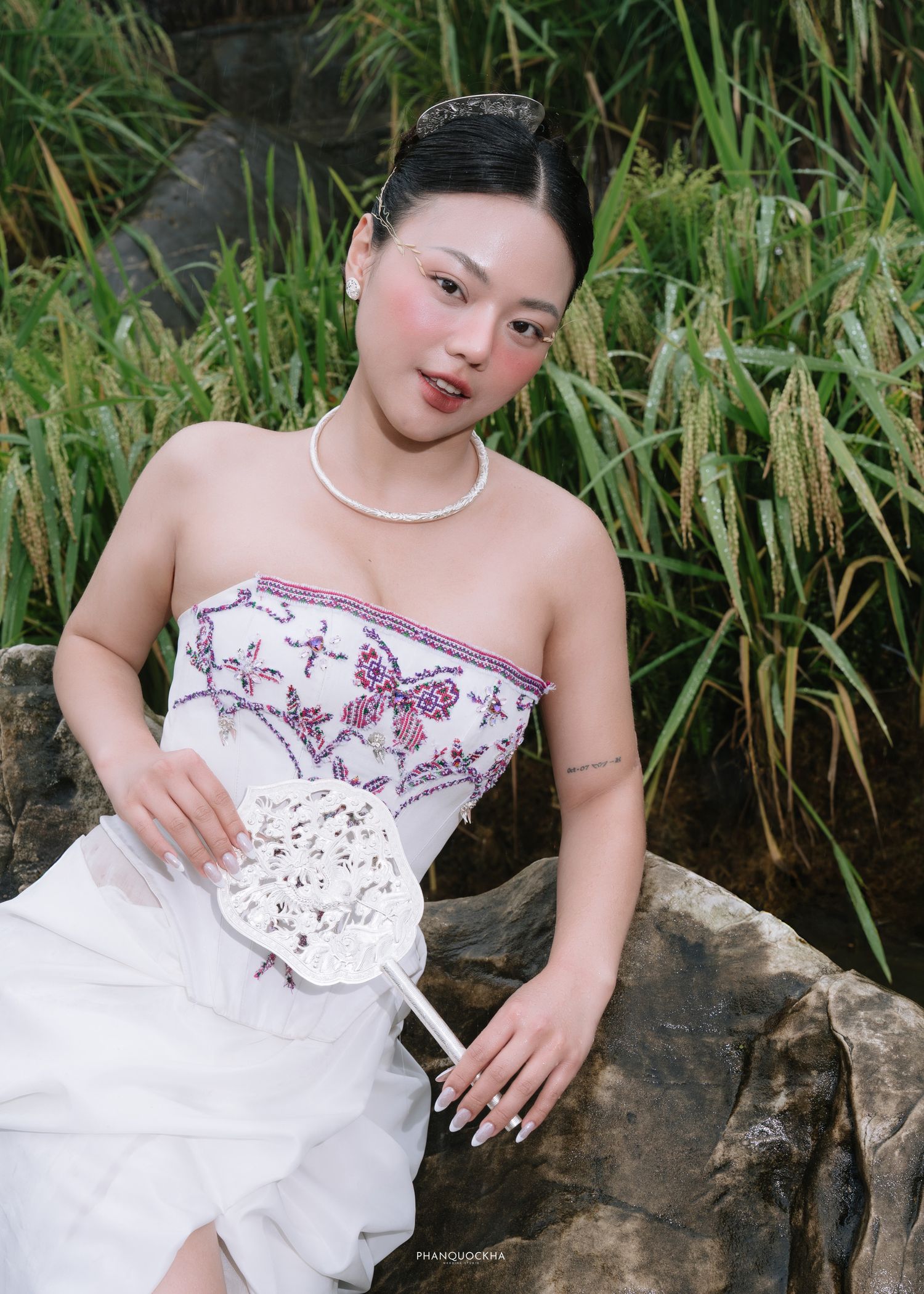 Young bride in strapless embroidered white gown holds bouquet while seated on rocks near tall grass.