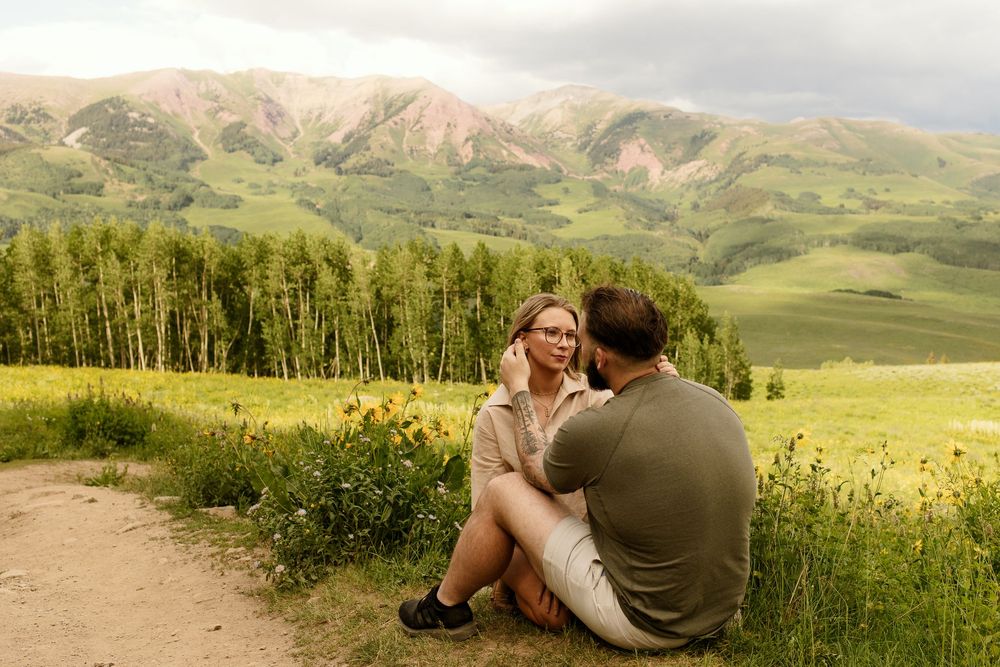 Two people sit together on a dirt path overlooking mountain meadows with aspen groves and rolling hills in Colorado.