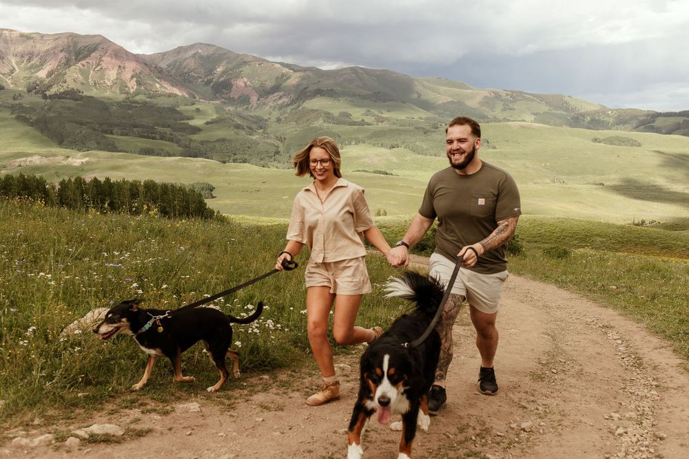 Hikers walk along a scenic mountain trail with a dog while exploring rugged mountain landscapes in summer weather.