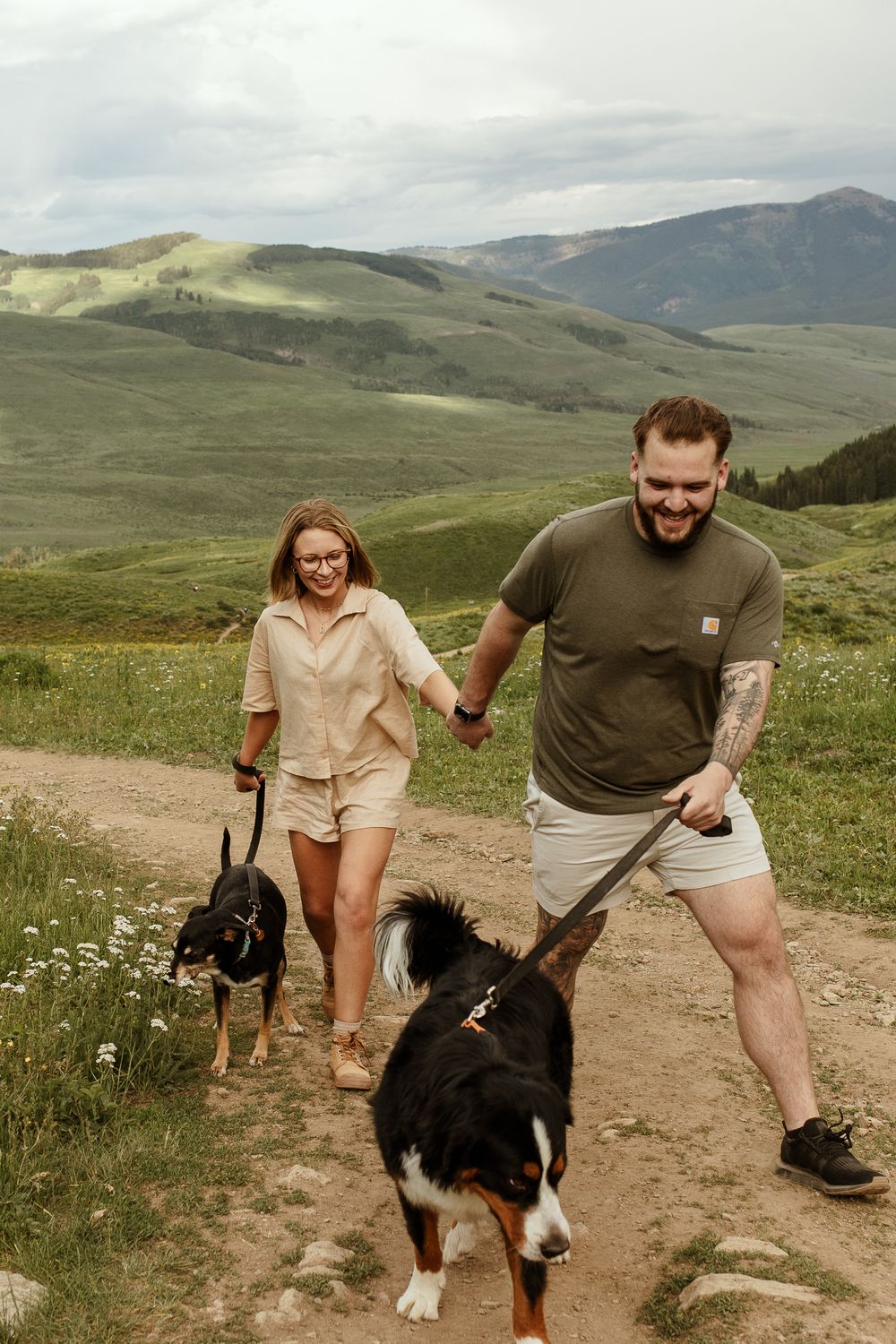 A couple and their Bernese Mountain Dog enjoy a scenic hike together on a mountain trail with rolling green hills.