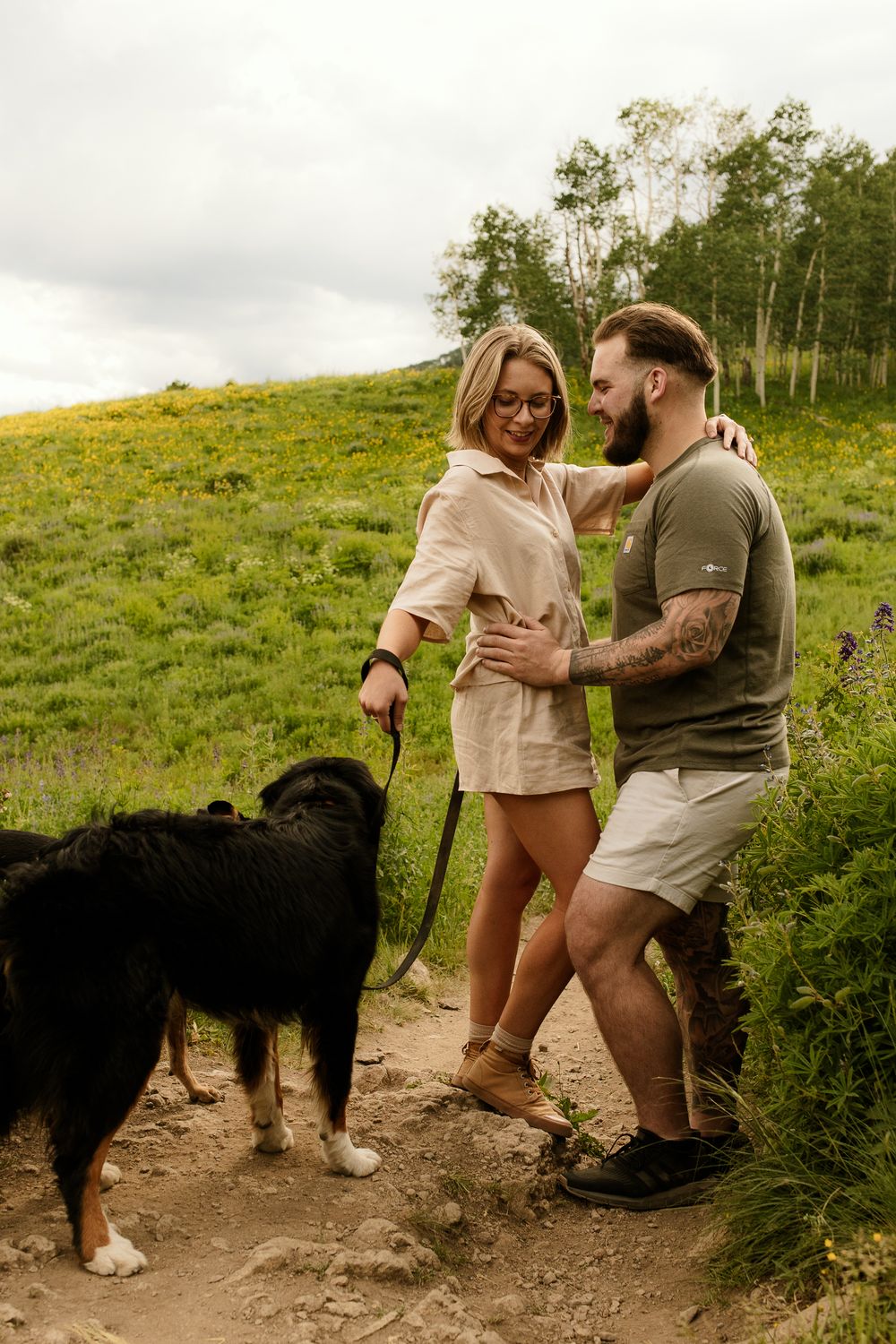 A couple shares a playful moment on a hiking trail with their black dog against a backdrop of lush mountain scenery.