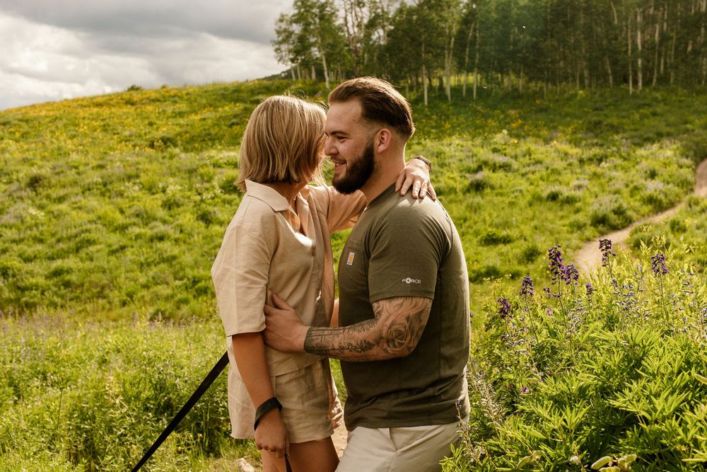 A romantic couple shares a tender moment in a sunlit meadow surrounded by wildflowers and green hills.