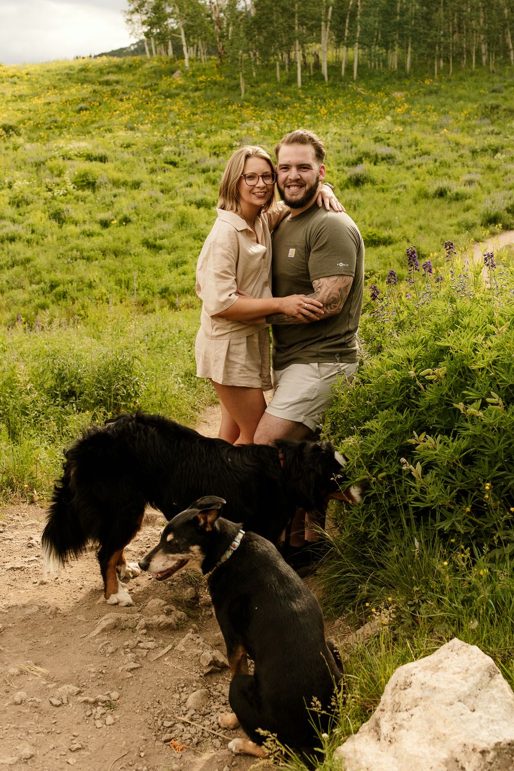 Couple embraces outdoors in natural setting with their black dog against scenic mountain landscape backdrop.