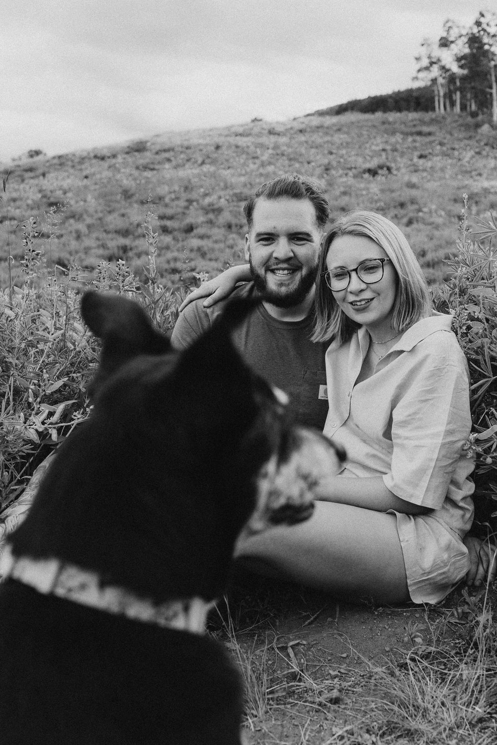 Couple lying in green grass field while their black dog photobombs the romantic moment in a series of candid shots.