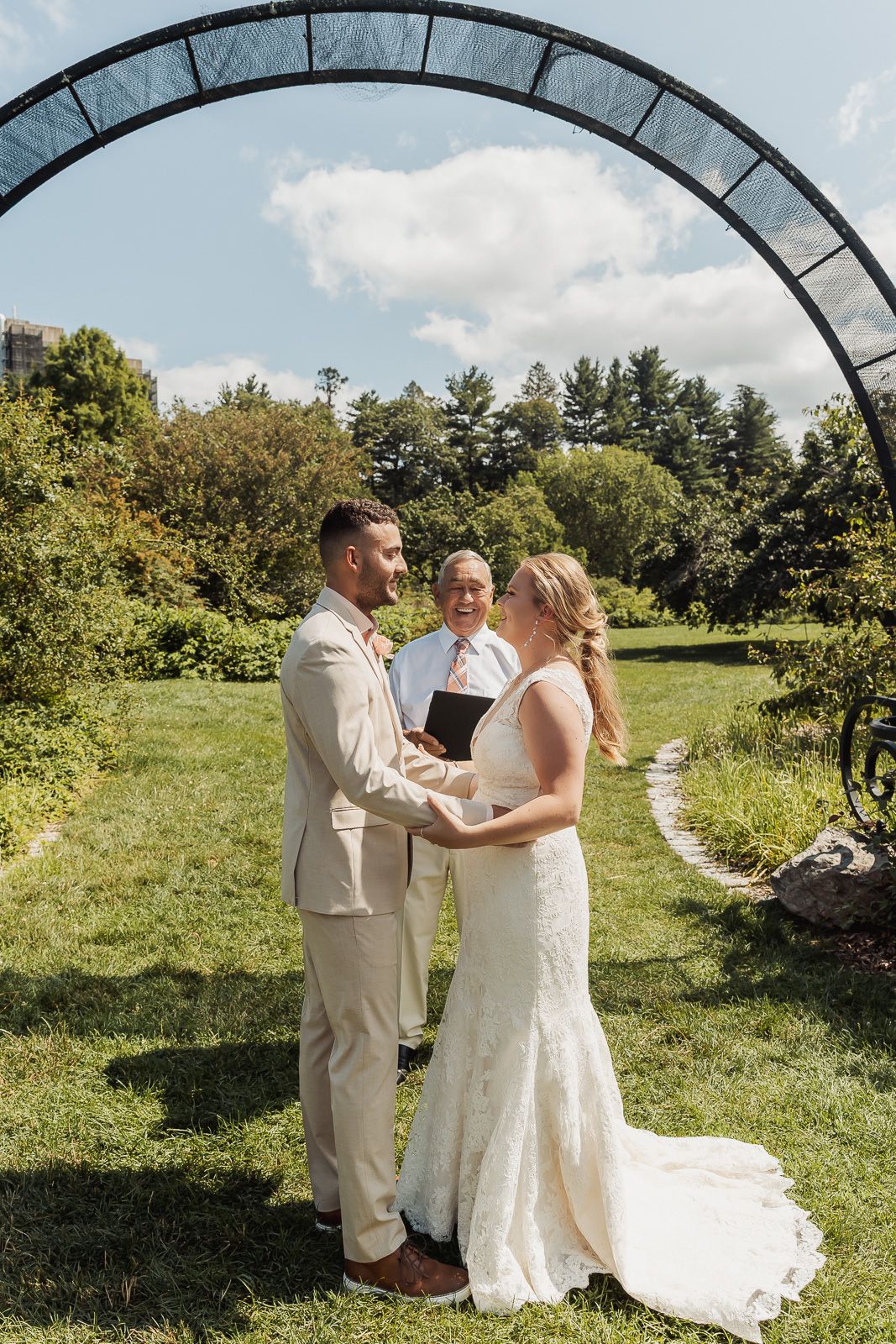 Wedding ceremony taking place under a rustic metal arch in a sunny garden setting on a beautiful summer day.
