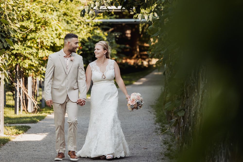 A couple in wedding attire walks along a sunlit tree-lined path during their outdoor wedding ceremony.