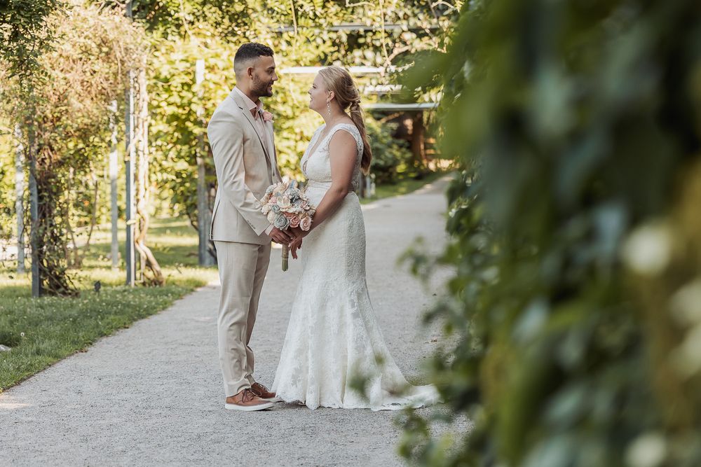 A couple shares a romantic moment in a sunlit garden path surrounded by lush greenery and natural light.