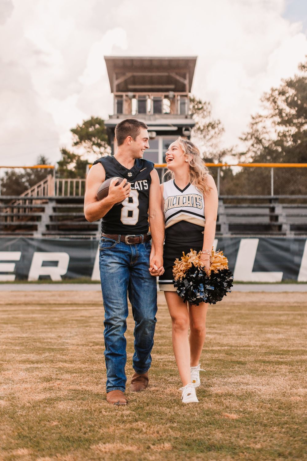 Couple in cheerleader and football uniforms walking on field together during romantic sunset photo shoot.