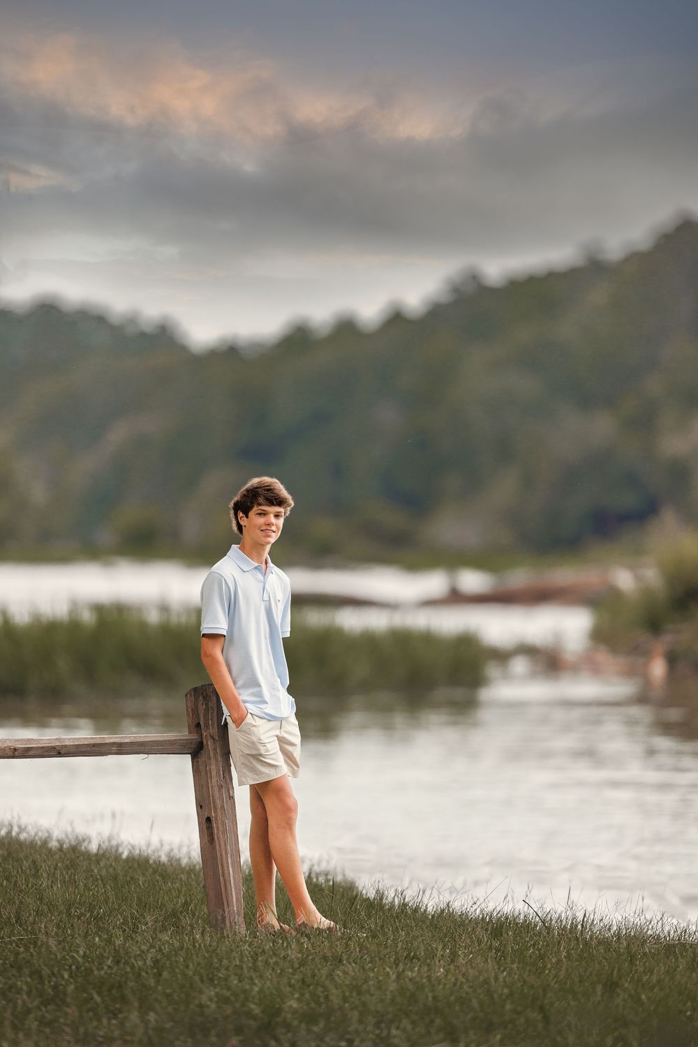 Young person in casual summer attire leans against wooden post by tranquil lakeside during sunset hour.