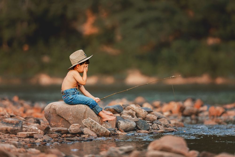 A child wearing a cowboy hat sits peacefully on rocks by a serene riverside in warm evening light.