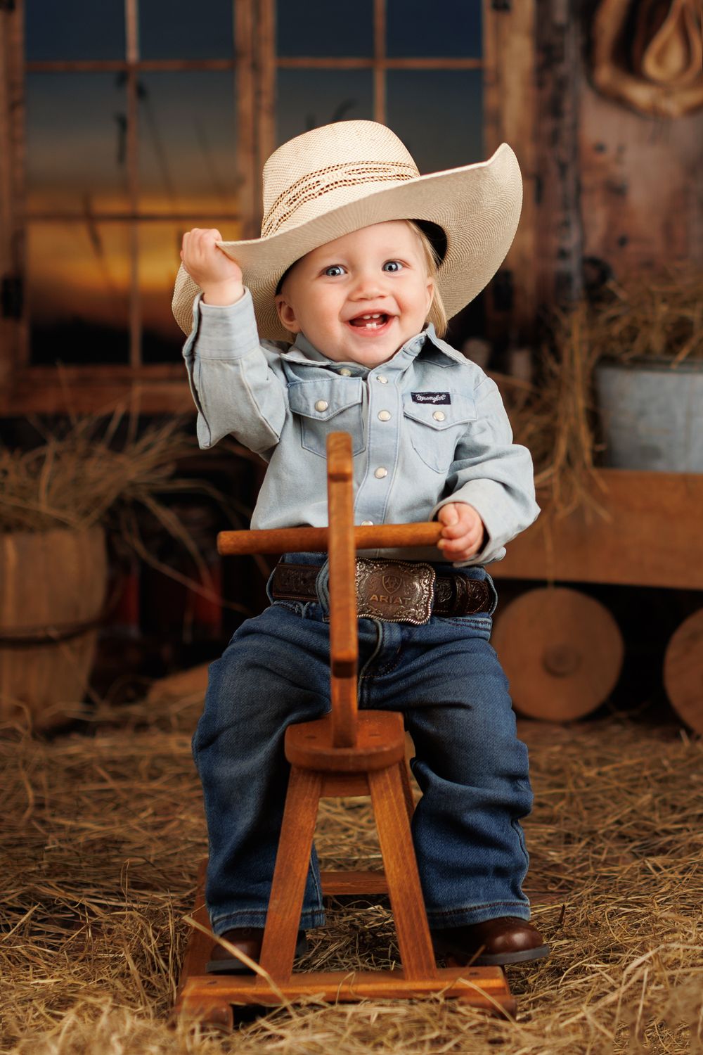 Country western styled infant sits on wooden stool in barn setting surrounded by hay.