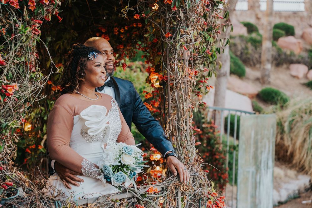 Couple in elegant wedding attire embracing outdoors next to ivy-covered walls and autumn foliage.