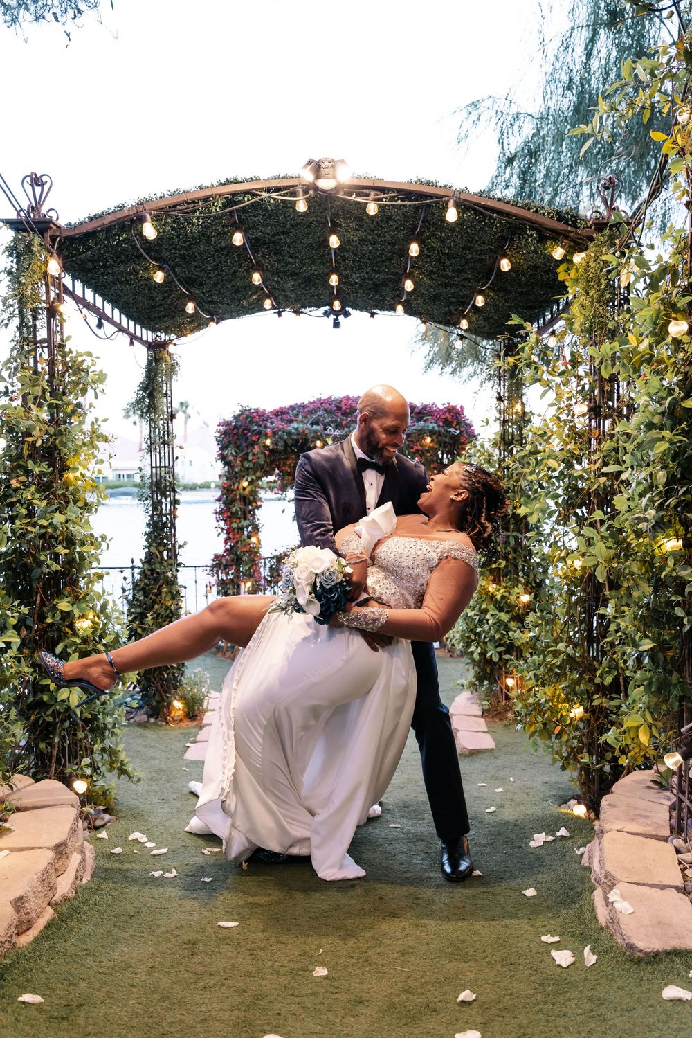 A couple shares a romantic dip kiss while dancing under a garden trellis decorated with string lights and pink flowers at sunset.