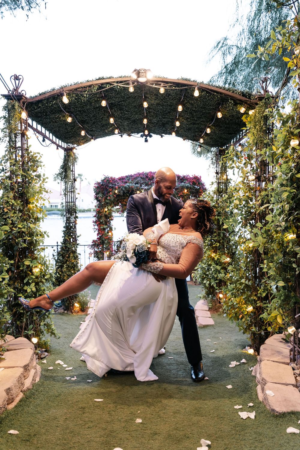 A romantic wedding couple dances at an outdoor garden venue decorated with string lights and floral arrangements.